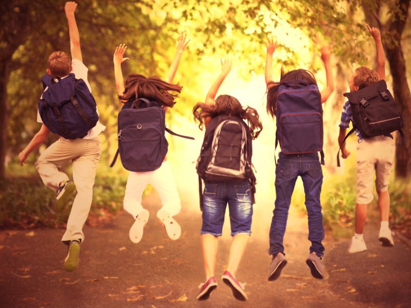 Rear-view of five children jumping in the air wearing backpacks